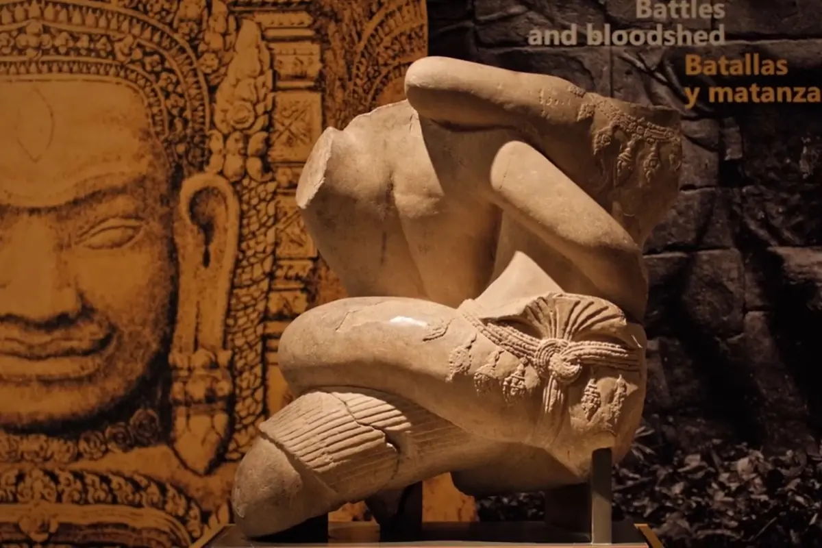 A stone sculpture of two Khmer grappling figures, one folded over the other, displayed in front of a large carved face at an Angkor exhibit