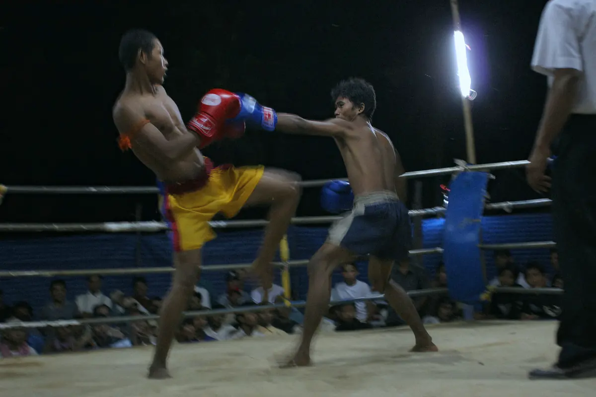 Two Kun Khmer fighters in gloves trade a knee and a straight punch in a floodlit ring while spectators watch from behind the ropes