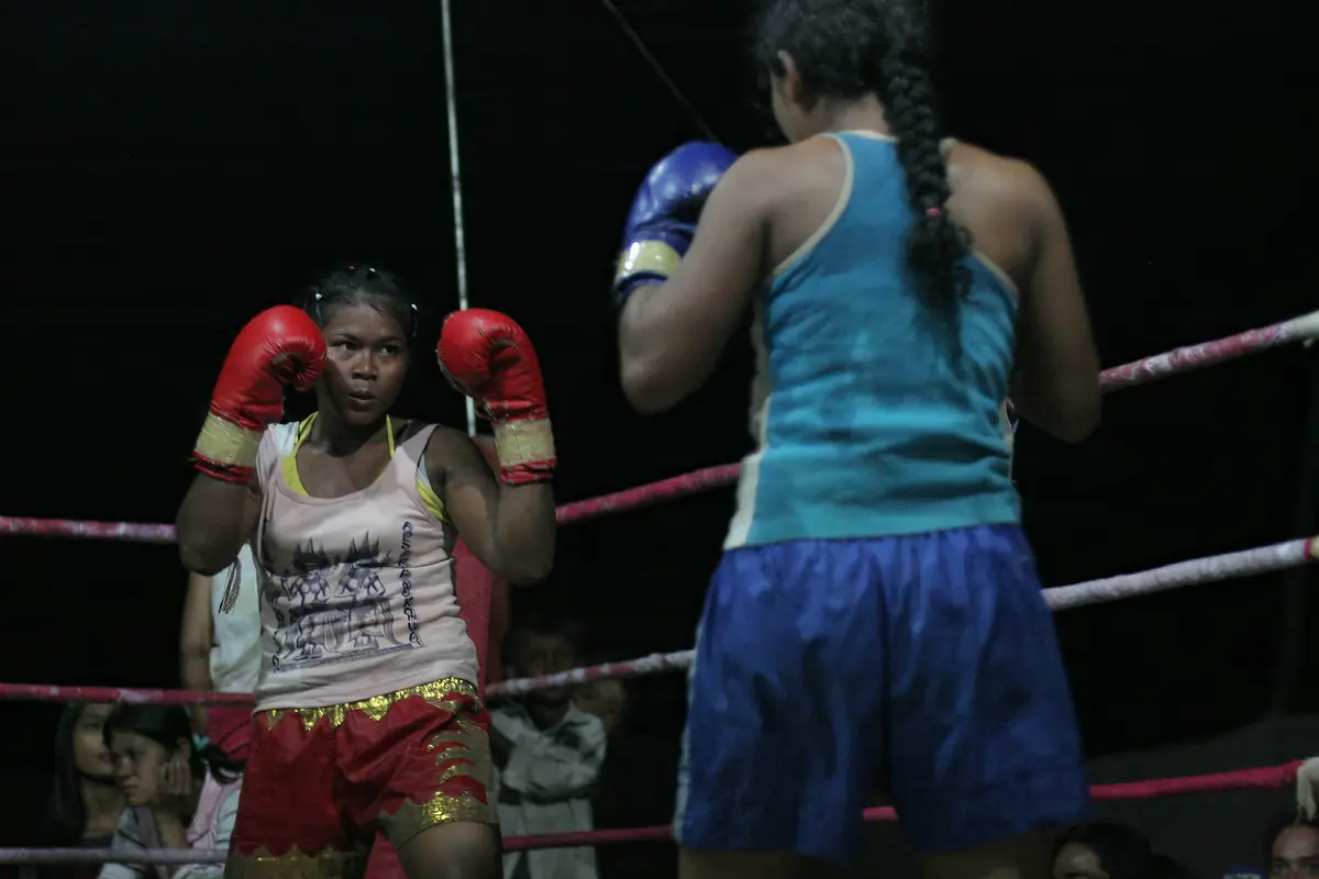 Two fighters in gloves face each other in a roped ring at night, one in a printed vest and red shorts with gold trim, the other in a blue vest with a plaited ponytail.