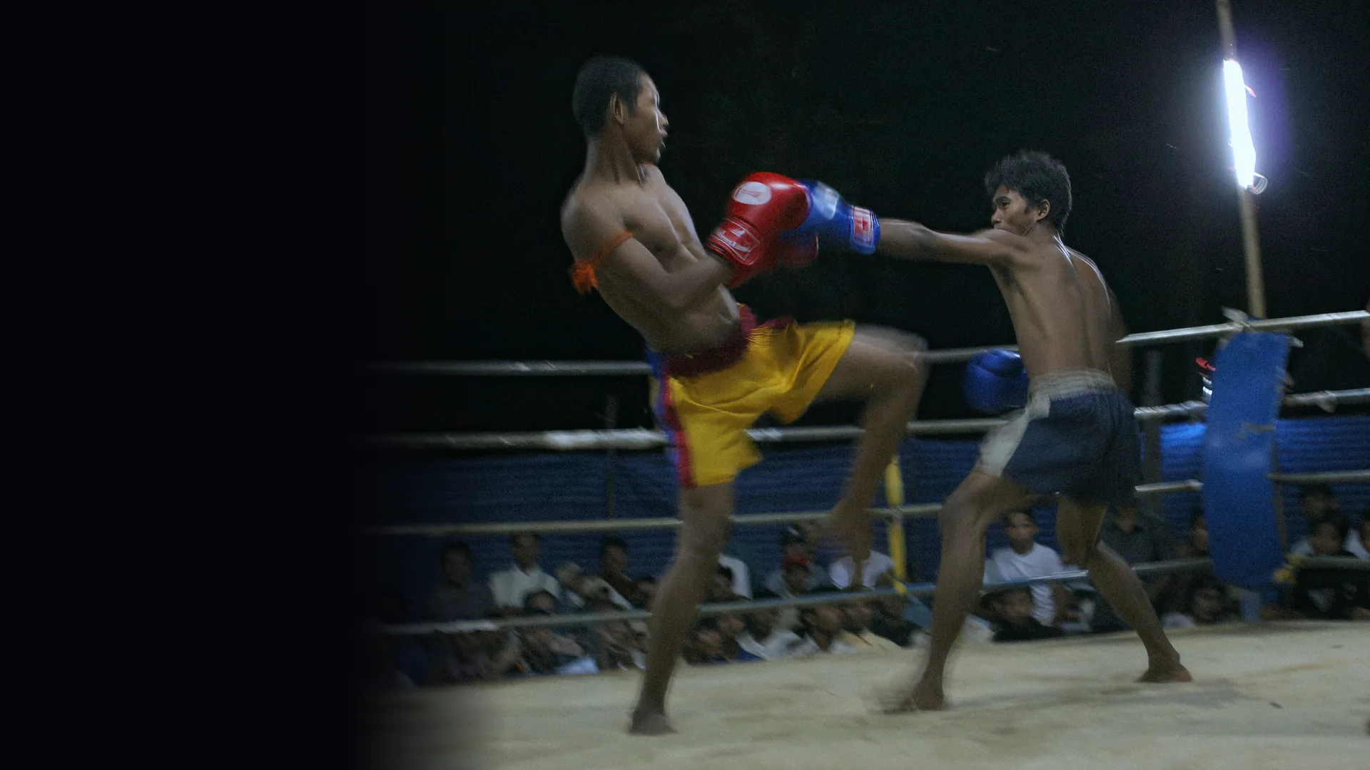 Two Kun Khmer fighters in gloves trade a knee and a straight punch in a floodlit ring, with spectators watching from behind the ropes.